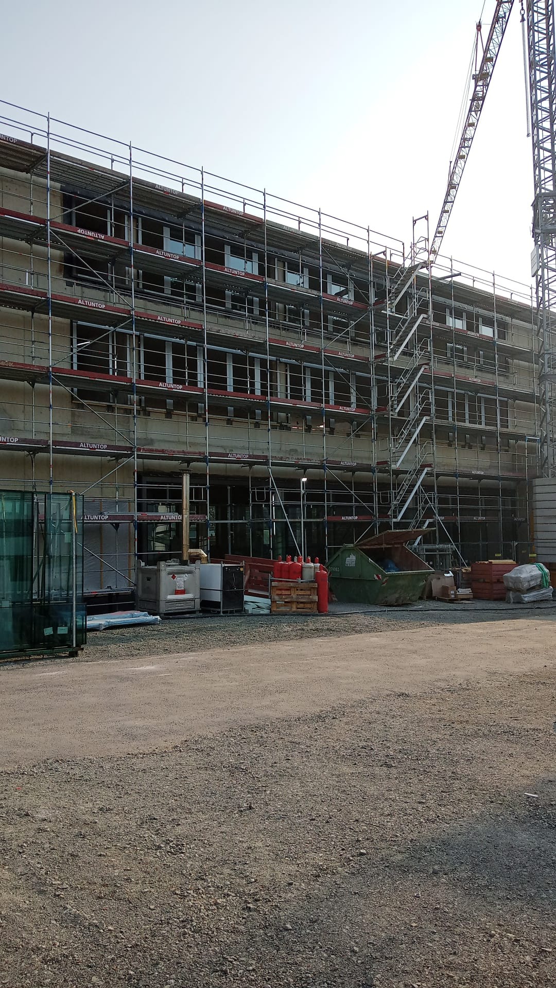 Budynek w trakcie montażu stolarki okiennej, widoczne rusztowania z napisem ALTUNTOP, dźwig budowlany w tle, teren budowy z gruzem i materiałami budowlanymi.