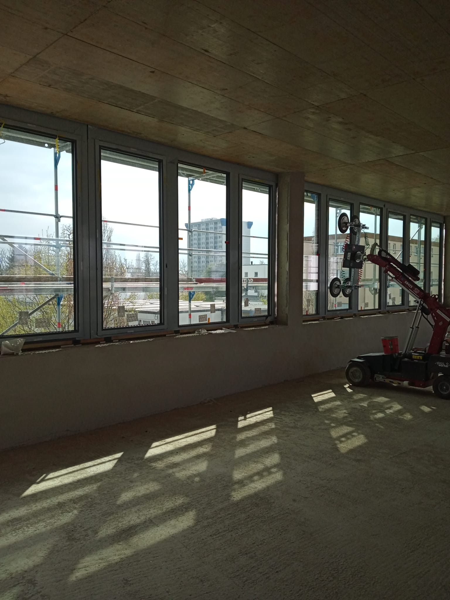 Montaż okien w surowym stanie budynku, widoczny podnośnik do szyb z przyssawkami, rusztowania za oknami i surowy betonowy strop.