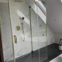 Kabina prysznicowa ze złotymi okuciami w łazience na poddaszu, marmurowe ściany i czarna podłoga, roślina doniczkowa w rogu.