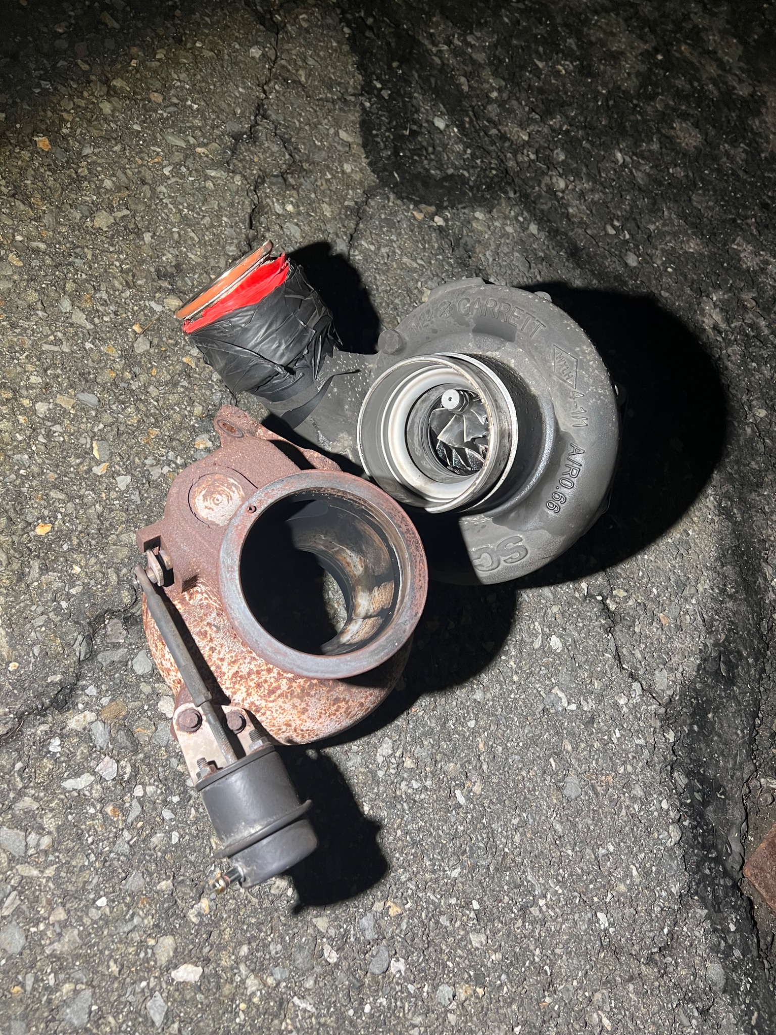 Używana turbosprężarka Garrett leżąca na asfalcie, z widocznymi śladami korozji i elementem zabezpieczonym czarną taśmą izolacyjną z czerwonym elementem wewnątrz.