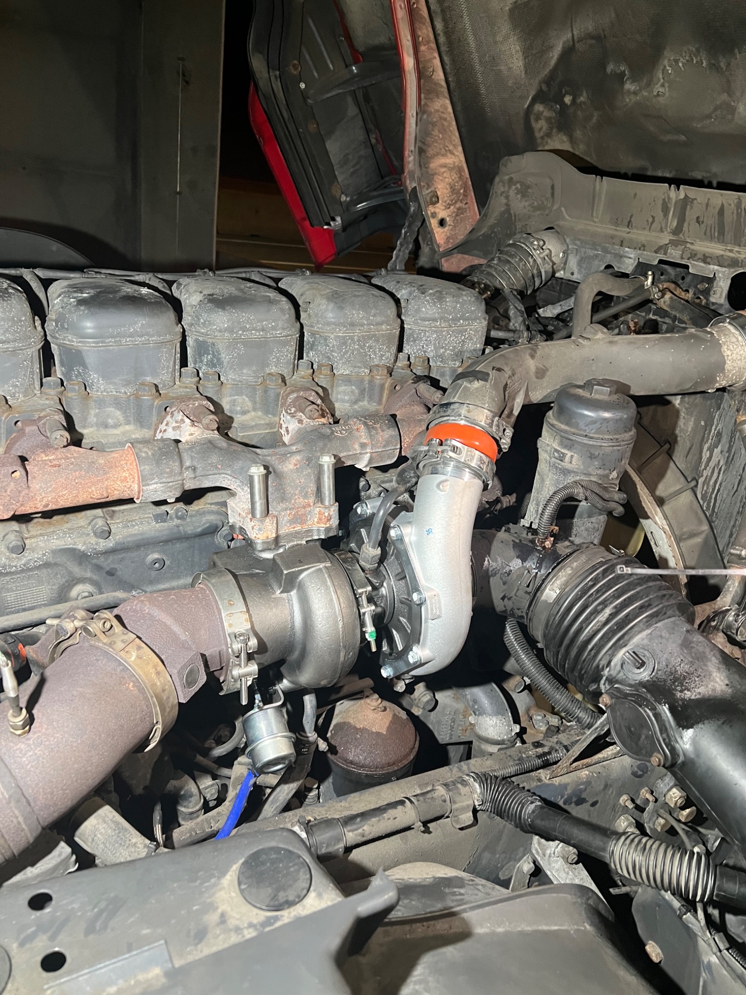 Silnik ciężarówki z nową turbosprężarką, widoczne zardzewiałe elementy układu wydechowego i świeżo zamontowana, srebrna turbosprężarka z niebieskim przewodem podciśnienia.