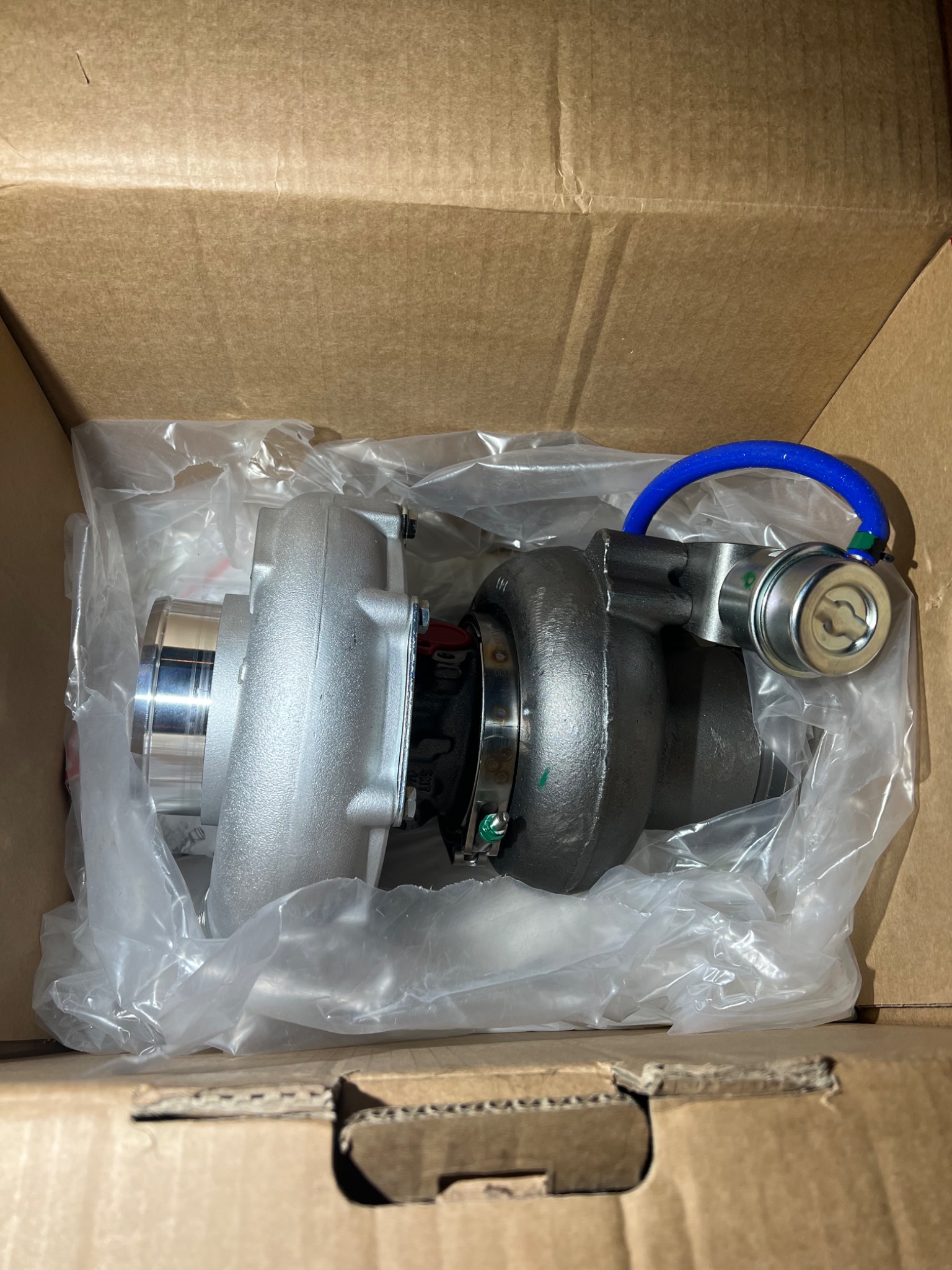 Nowa turbosprężarka w pudełku, zapakowana w folię, z widocznym niebieskim przewodem.