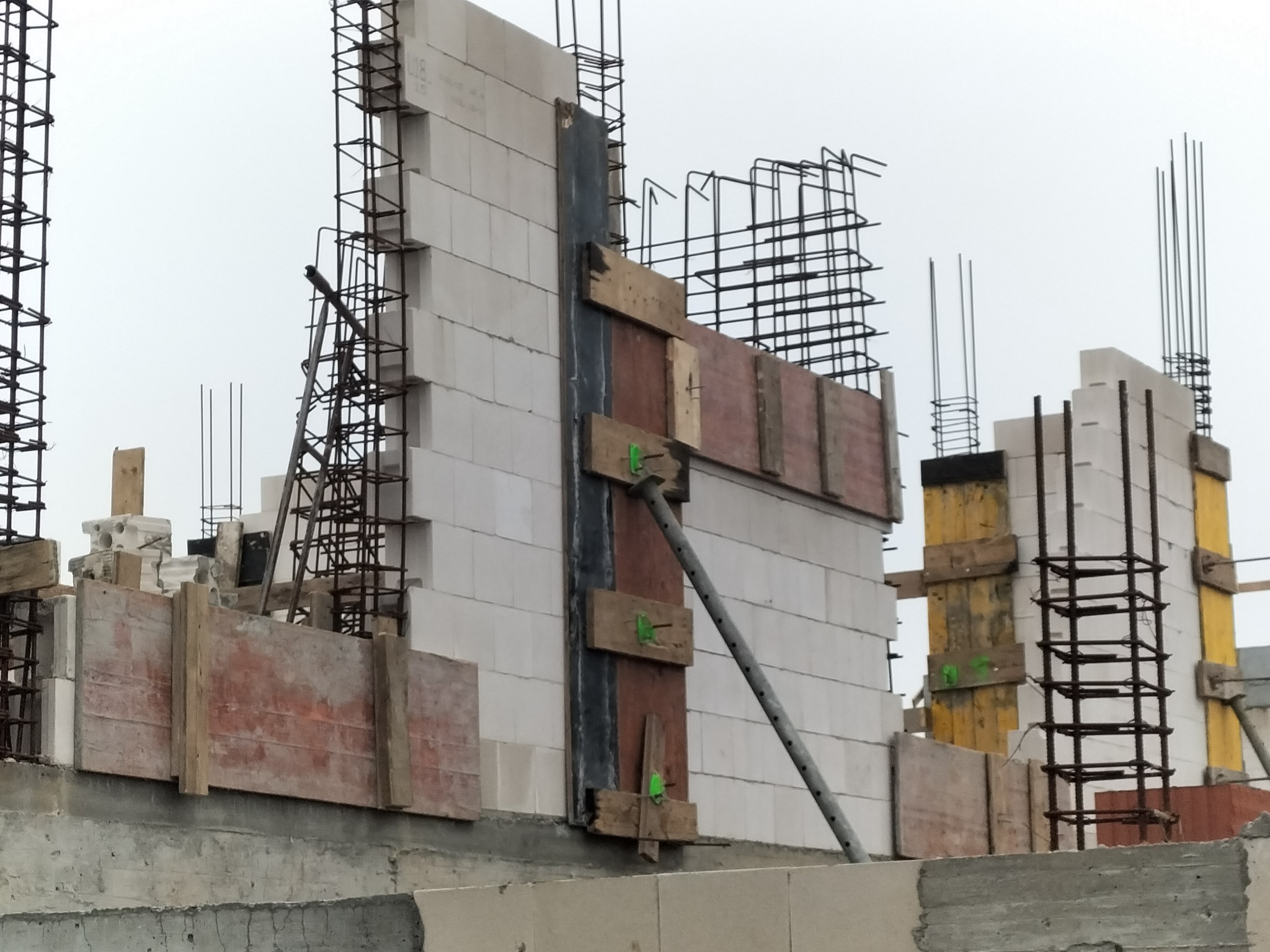 Wznoszenie ściany z bloczków betonu komórkowego z widocznym zbrojeniem i szalunkiem drewnianym, wspartym metalową podporą, na surowym fundamencie.