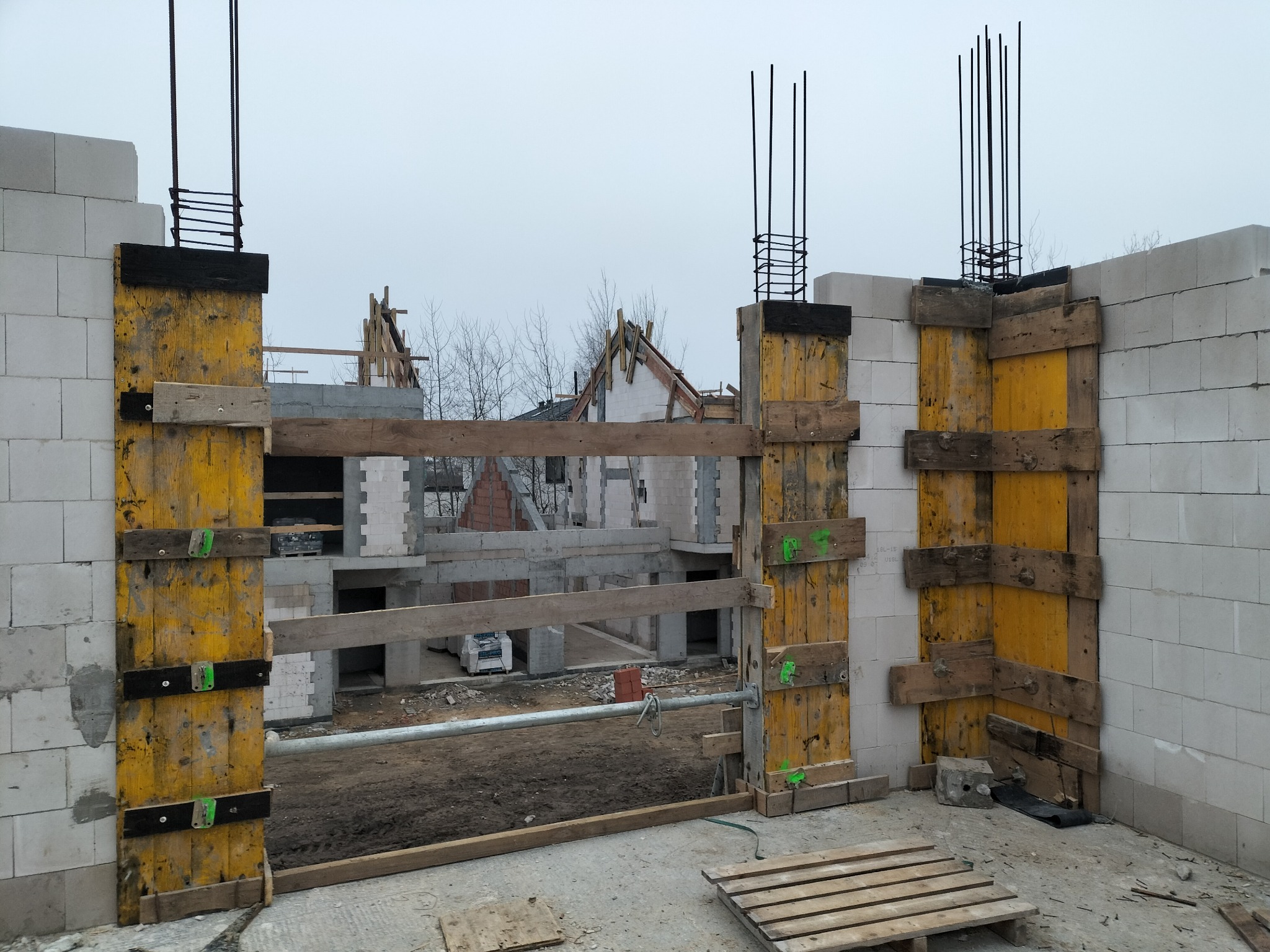 Budowa domu jednorodzinnego z bloczków, widoczne szalunki i zbrojenie pionowe, w tle konstrukcja dachu sąsiedniego budynku.