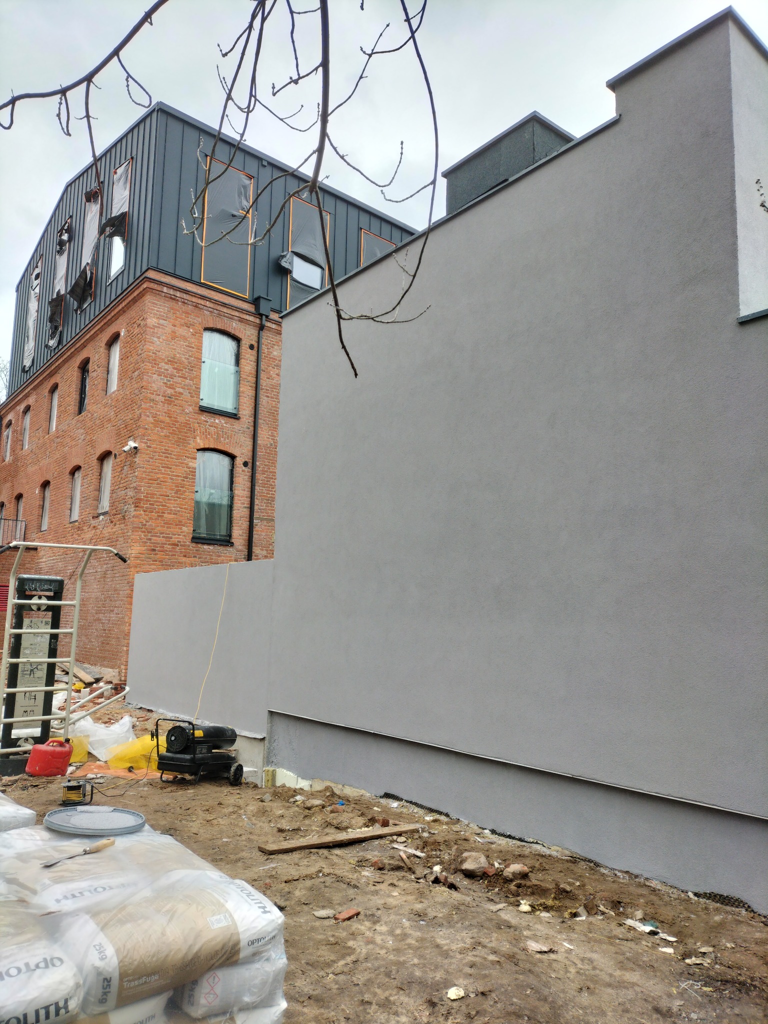Świeżo otynkowana szara ściana budynku z widocznymi workami zaprawy, nagrzewnicą i narzędziami na placu budowy, w tle budynek z cegły i blachy trapezowej.