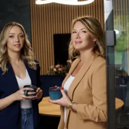 Dwie kobiety w biurze, trzymające identyczne, ceramiczne kubki z niebieskim gradientem, ubrane w eleganckie marynarki, z drewnianą ścianą w tle i nowoczesnym oświetleniem sufitowym.