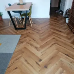 Jasne panele ułożone w jodełkę w salonie, widoczny fragment dywanu, stół z krzesłami i komoda w tle.