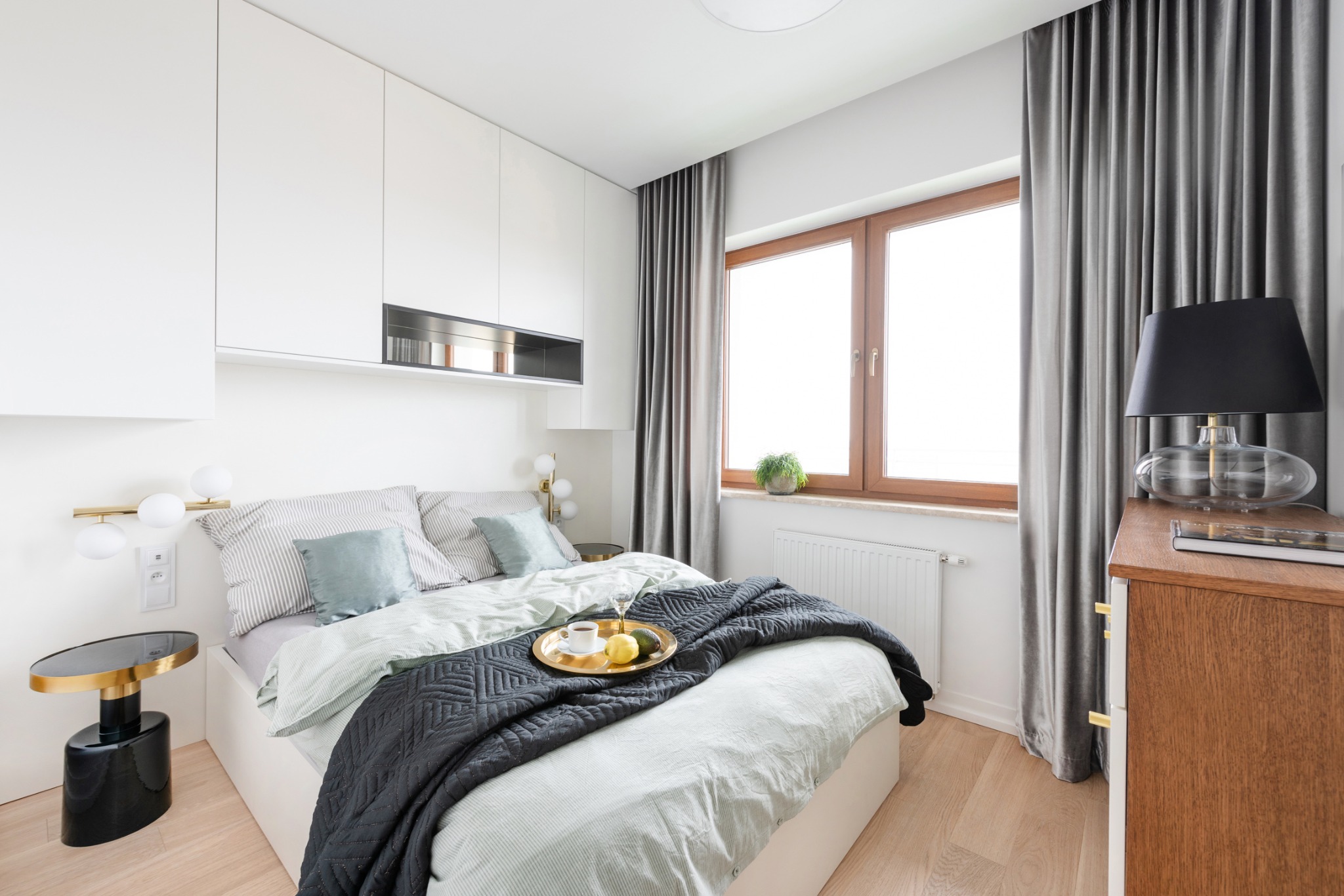 Jasna sypialnia z łóżkiem, stolikiem nocnym i zasłonami; na łóżku taca ze śniadaniem.