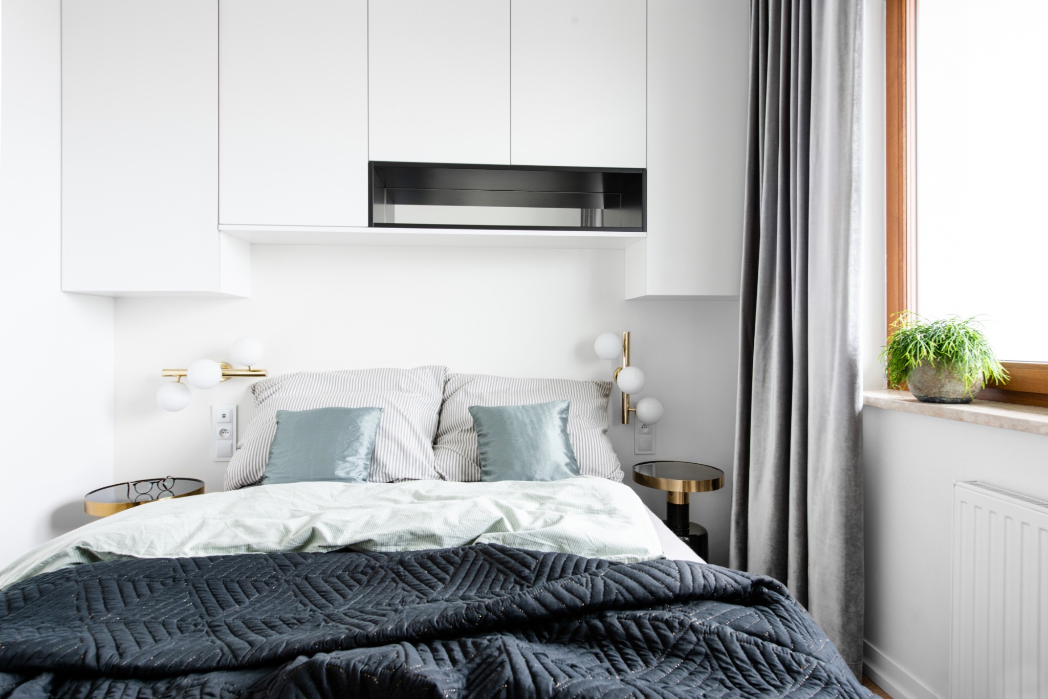 Nowoczesna sypialnia: czarna pikowana narzuta, pastelowa pościel, lampki nocne z kulistymi kloszami, szafki wiszące nad łóżkiem, okno z zielonym kwiatem doniczkowym.