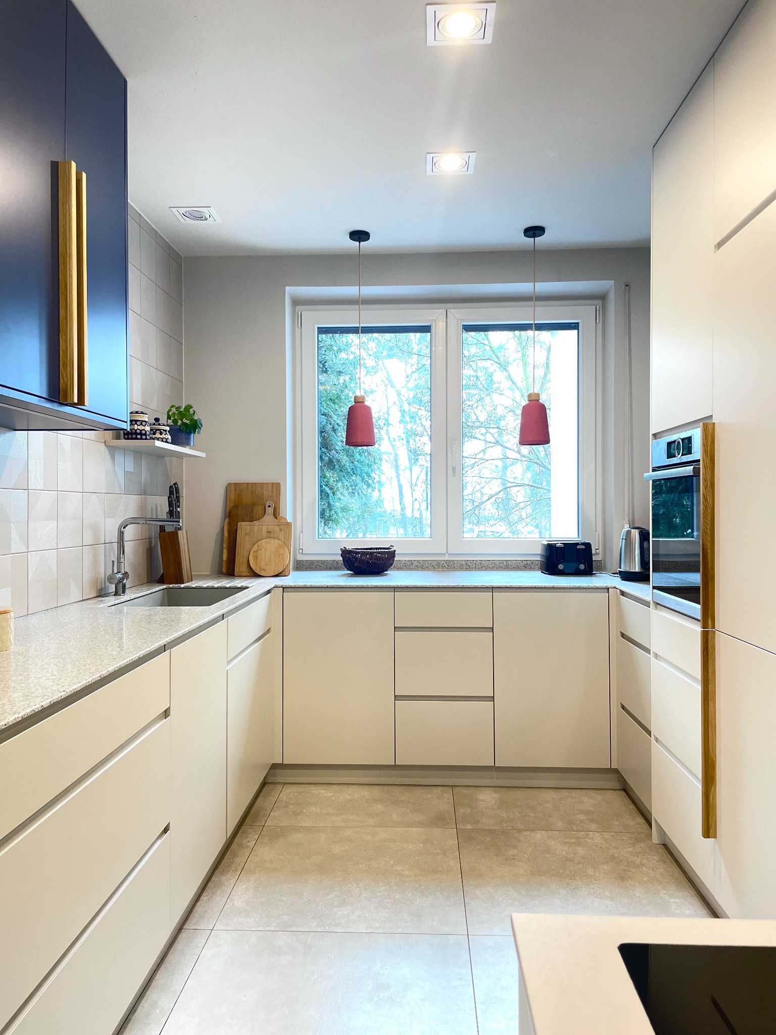 Nowoczesna kuchnia w odcieniach bieli i granatu z drewnianymi akcentami, wyposażona w granitowy blat i podwieszane lampy w kolorze bordo, z widokiem na drzewa za oknem.
