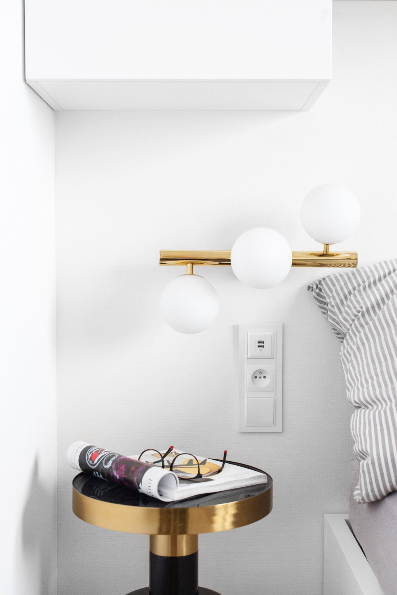 Nowoczesna sypialnia z okrągłym, czarno-złotym stolikiem nocnym, na którym leżą okulary i zwinięta gazeta, oświetlona złotą lampą ścienną z trzema białymi kulami, obok łóżka z szarą pościelą w paski.
