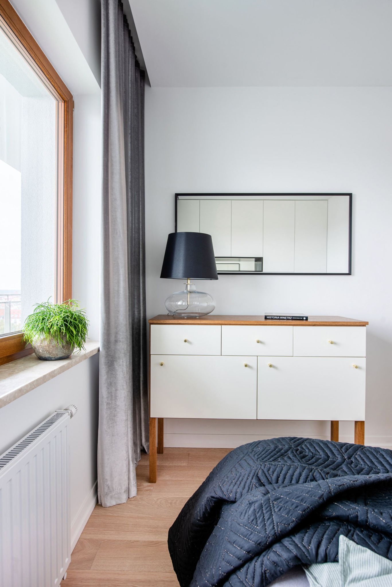 Jasna sypialnia z drewnianą komodą, lustrem w czarnej ramie, lampą z czarnym abażurem i rośliną doniczkową na parapecie okna.