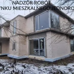 Budynek mieszkalny jednorodzinny w trakcie budowy, widoczne surowe ściany z bloczków, zamontowane okna, ośnieżone otoczenie i drzewa bez liści w zimowej scenerii. W prawym dolnym rogu widoczne logo...