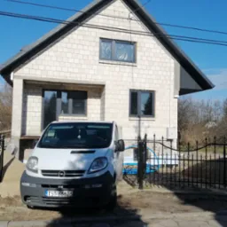 Surowa bryła dwukondygnacyjnego domu z białego bloczka, z zamontowanymi oknami o grafitowych ramach, częściowo ocieplona, z zaparkowanym białym vanem Opel Vivaro na posesji. W tle widoczne drzewa...