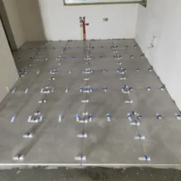 Szare płytki podłogowe w trakcie układania w surowym wnętrzu, z widocznymi systemami poziomowania płytek i instalacją hydrauliczną na ścianie.