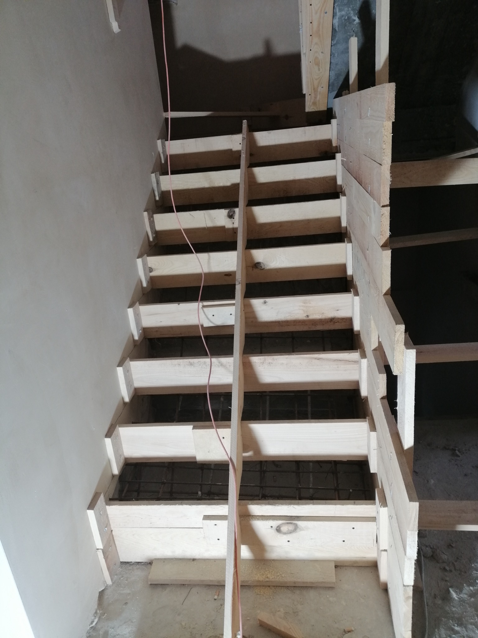 Surowe drewniane schody w trakcie budowy, widoczne elementy konstrukcyjne i zbrojenie, kabel zwisający z góry, surowe ściany w tle.