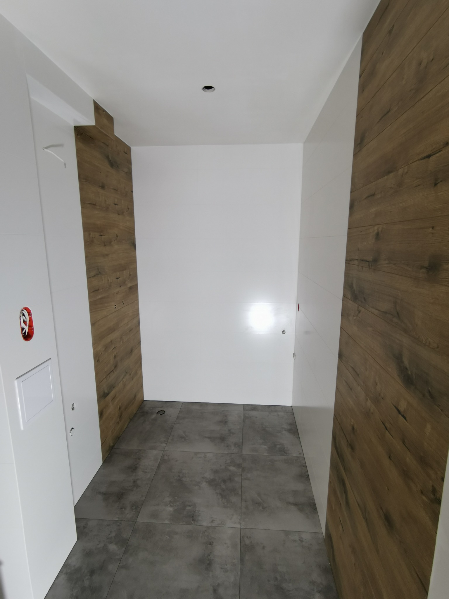 Wnętrze łazienki w trakcie remontu: szare płytki podłogowe, częściowo wykończone ściany z imitacją drewna, białe ściany i drzwi z widocznymi puszkami elektrycznymi.