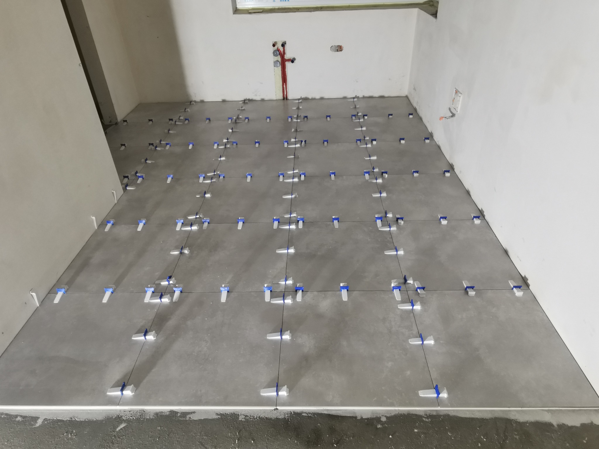 Szare płytki podłogowe w trakcie układania w surowym wnętrzu, z widocznymi systemami poziomowania płytek i instalacją hydrauliczną na ścianie.