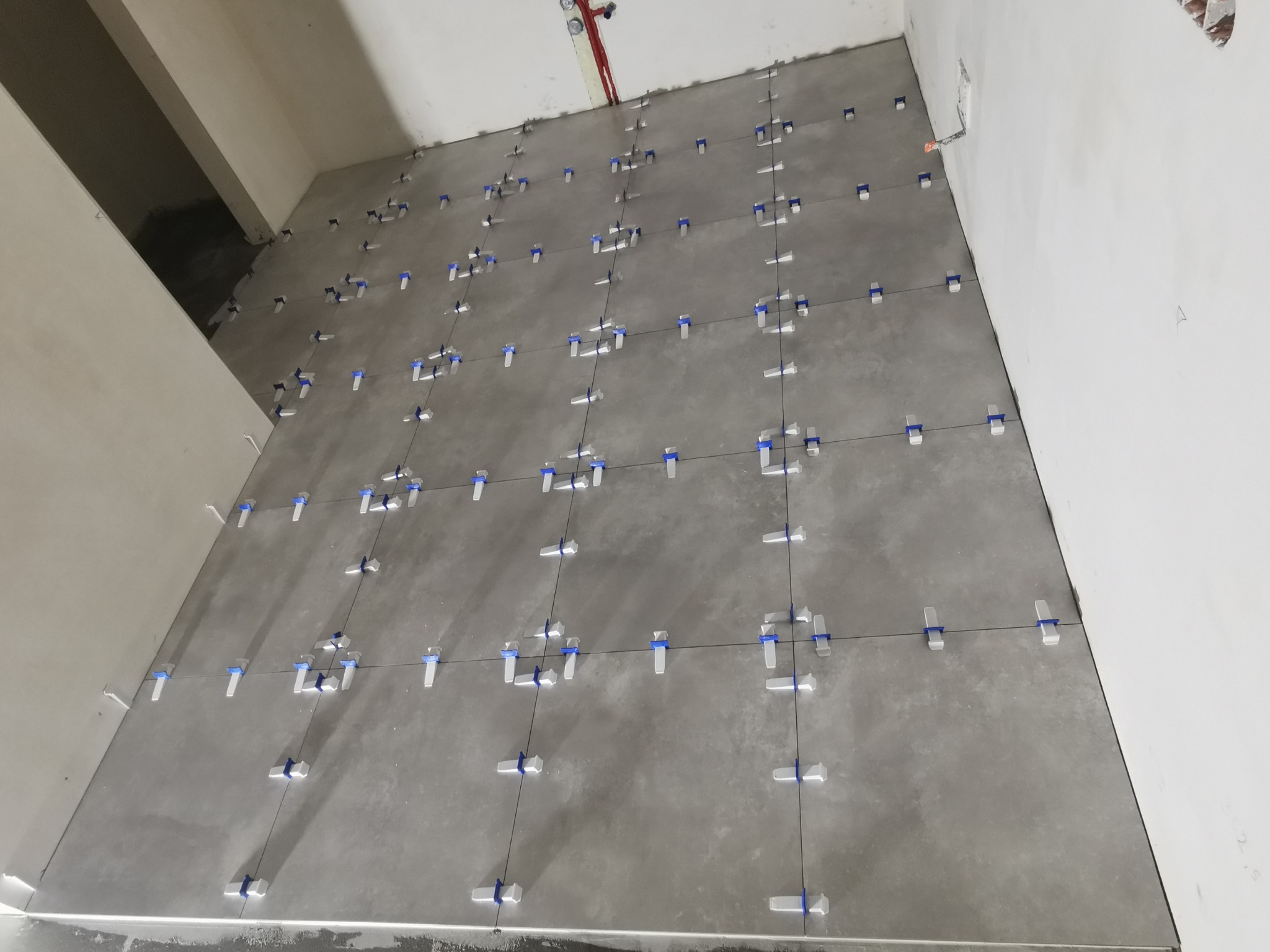 Szare płytki podłogowe w trakcie układania, z użyciem białych klinów i niebieskich krzyżyków dystansowych, widoczne przewody instalacji pod sufitem.