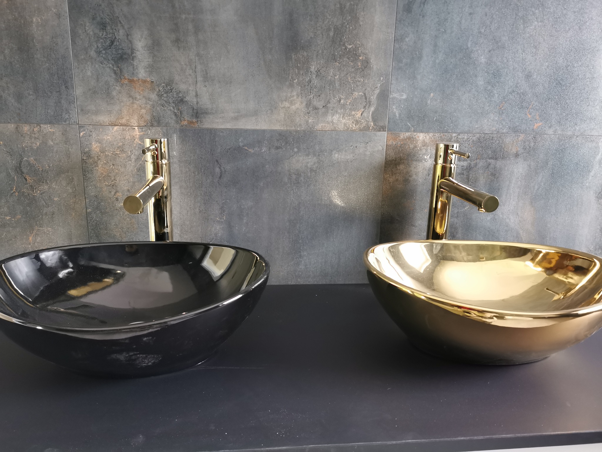 Dwie umywalki nablatowe: czarna i złota, z błyszczącymi złotymi kranami na tle szarej, kamiennej ściany z płytek.