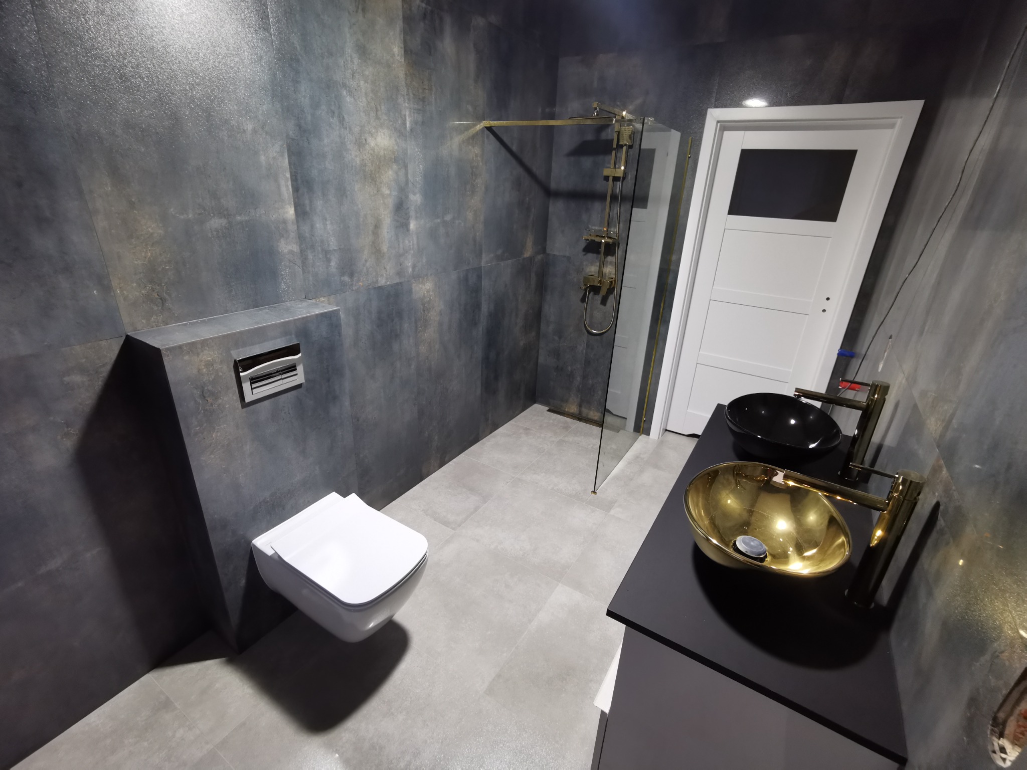Nowoczesna łazienka z szarymi płytkami imitującymi beton, wiszącą toaletą, złotą armaturą i umywalką, oraz kabiną prysznicową ze złotymi elementami.