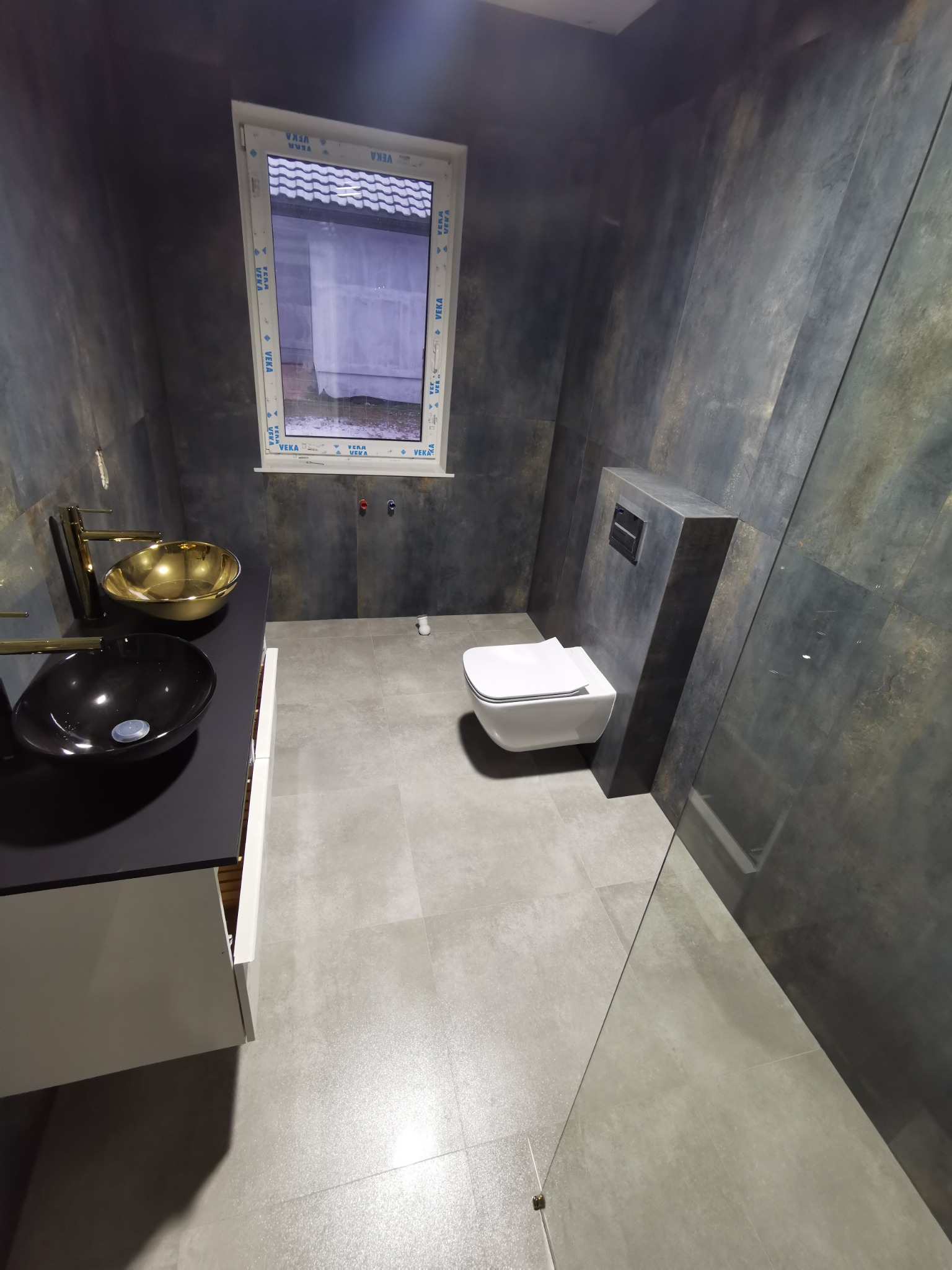 Nowoczesna łazienka z umywalkami w kolorze czarnym i złotym, wisząca toaleta, szare płytki na ścianach i podłodze, okno z widokiem na zewnątrz.