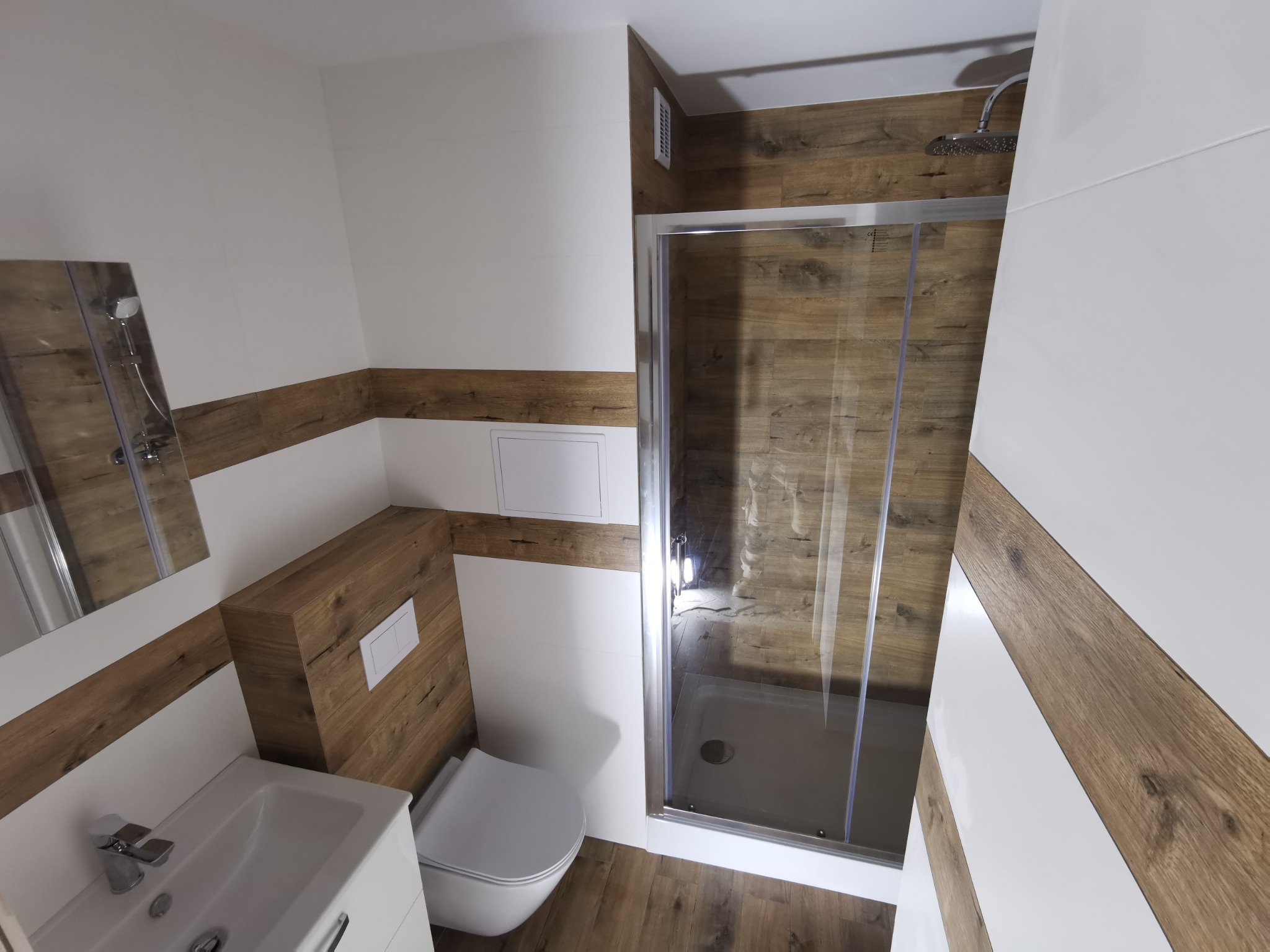 Nowoczesna łazienka z kabiną prysznicową typu walk-in, wykończona białymi i drewnopodobnymi płytkami, z umywalką i toaletą w minimalistycznym stylu.