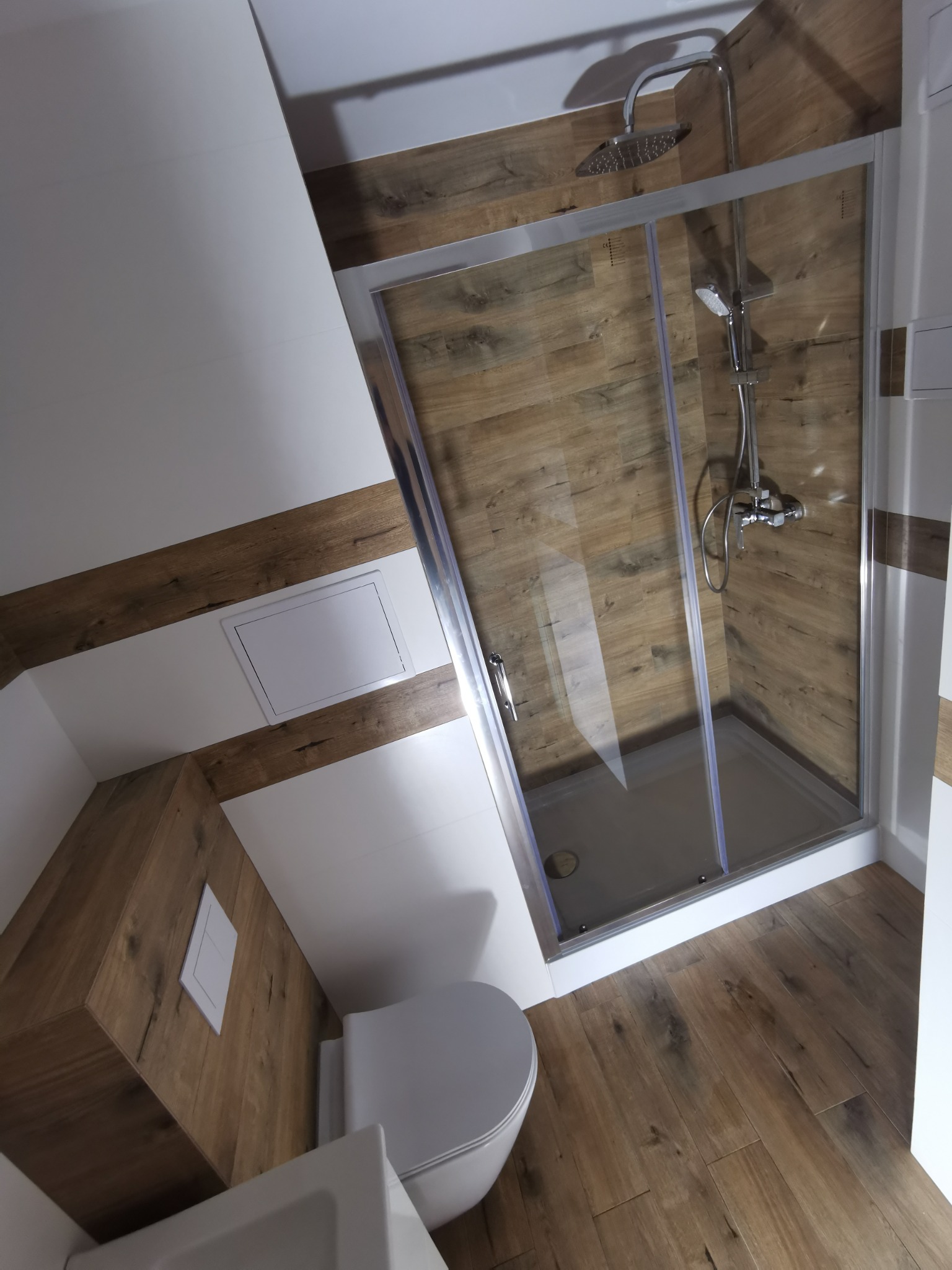 Ukośna łazienka z kabiną prysznicową typu walk-in, drewnopodobnymi płytkami na ścianach i podłodze, oraz białymi elementami wykończeniowymi.