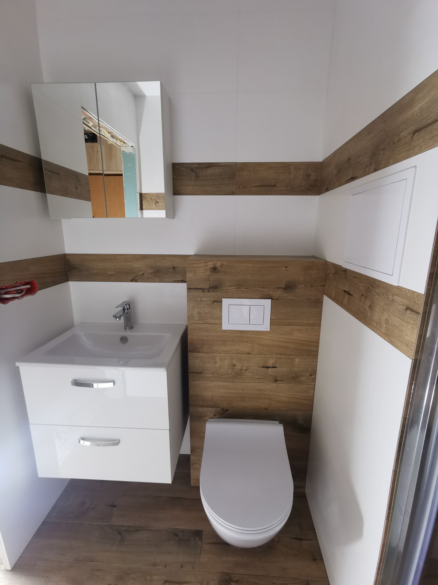 Nowoczesna łazienka po remoncie: biała umywalka z szafką, wisząca toaleta i pasy dekoracyjne imitujące drewno na ścianach, odbicie fragmentu innego pomieszczenia w lustrze szafki.