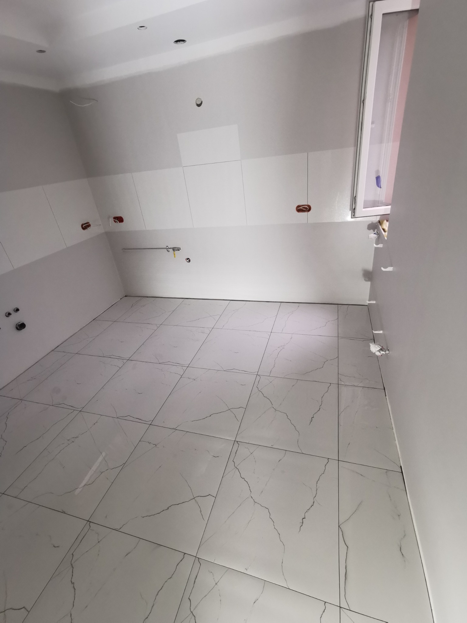 Wykończona łazienka w trakcie remontu: białe płytki imitujące marmur na podłodze, częściowo wykończone białe ściany z widocznymi instalacjami hydraulicznymi i elektrycznymi, okno z delikatną firanką.