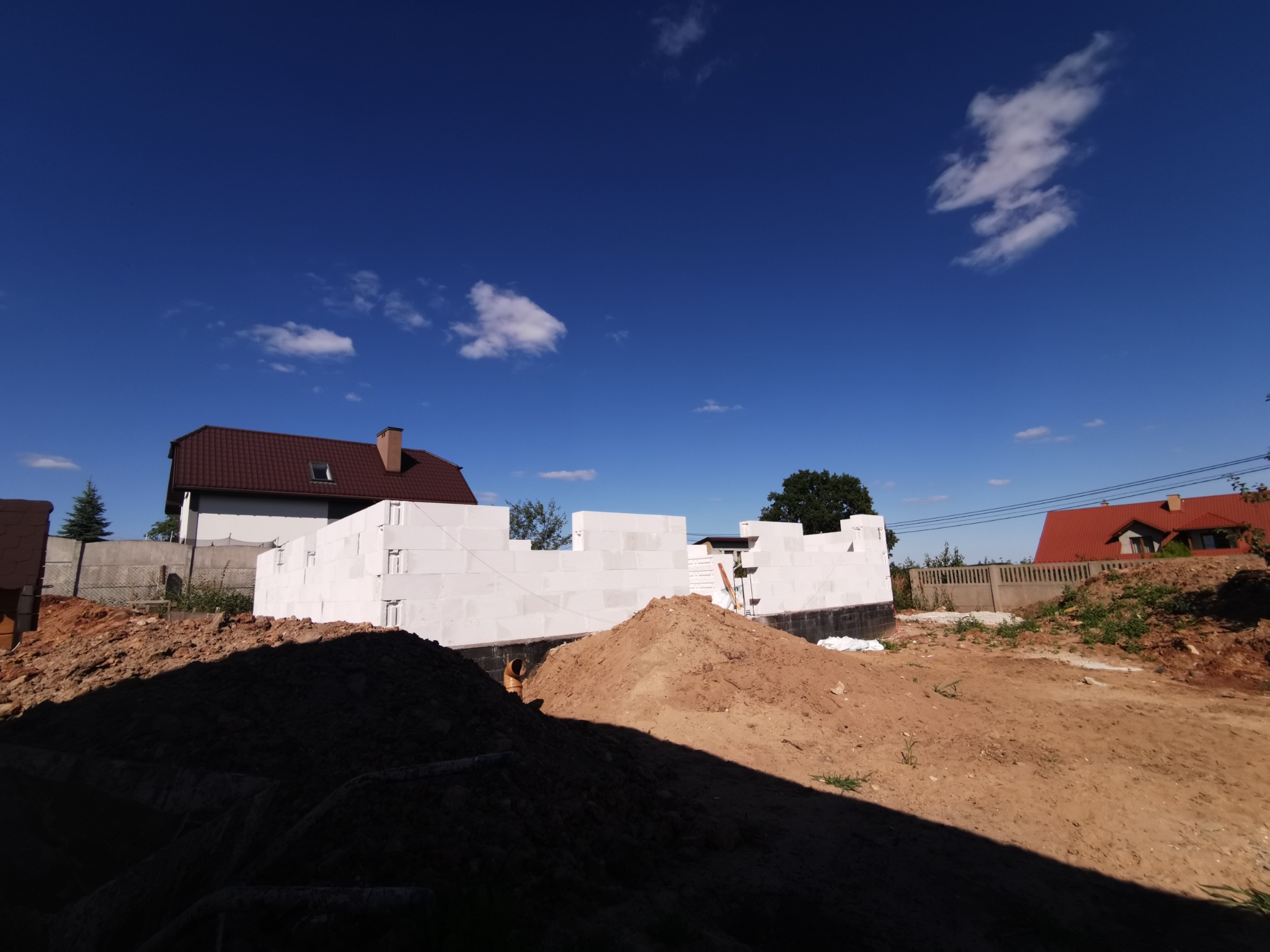 Wznoszone ściany parteru z białych bloczków betonu komórkowego na fundamencie z hydroizolacją, widok z poziomu gruntu na placu budowy w słoneczny dzień, w tle domy jednorodzinne.