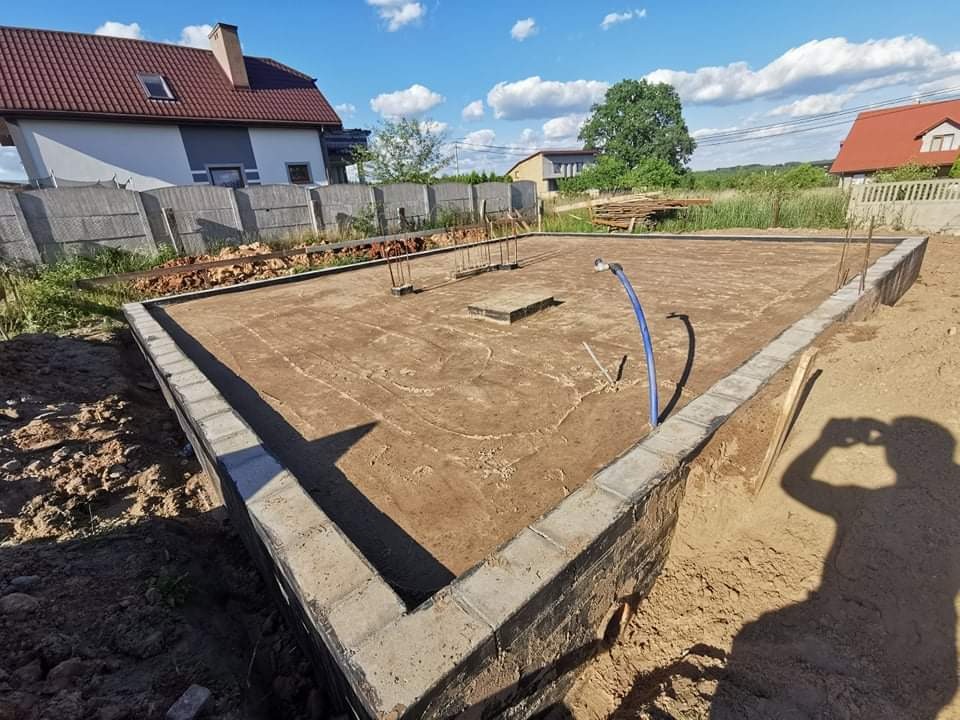 Wylany fundament pod budynek mieszkalny, widoczne wystające pręty zbrojeniowe i niebieski wąż doprowadzający wodę na plac budowy, w tle domy jednorodzinne i błękitne niebo.