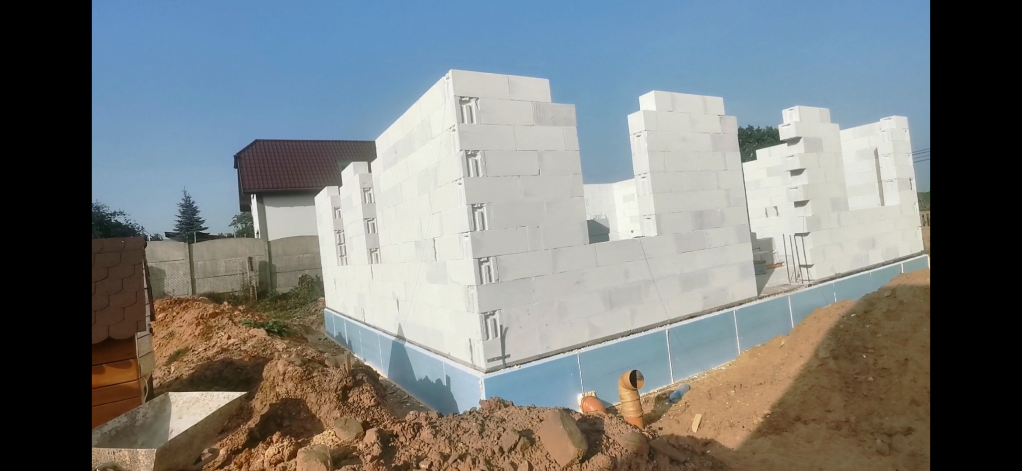 Wznoszenie murów domu z białych bloczków betonowych, widoczne fundamenty zabezpieczone izolacją, teren wokół w stanie surowym.