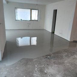 MB-BUD - Wylewka samopoziomująca w jasnym pokoju z oknem zabezpieczonym folią. Część podłogi jeszcze niezalana, widoczny surowy beton. Ściany przygotowane do malowania.