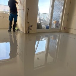 MB-BUD - Wylewka samopoziomująca w jasnym pomieszczeniu z dużym oknem balkonowym. Pracownik w butach z kolcami rozprowadza masę. Widoczne instalacje elektryczne w ścianach.