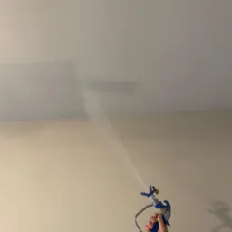 Natryskowe malowanie sufitu pistoletem w kolorze jasnego beżu, widoczna ręka w niebieskim ubraniu trzymająca urządzenie, cień pistoletu na ścianie.