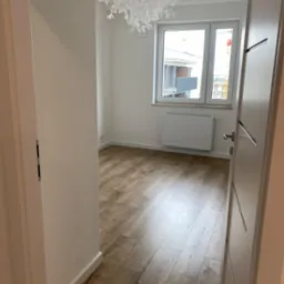 Pusty pokój z panelami podłogowymi w odcieniach brązu, białymi ścianami, grzejnikiem pod oknem i nowoczesną lampą sufitową w kształcie liści.