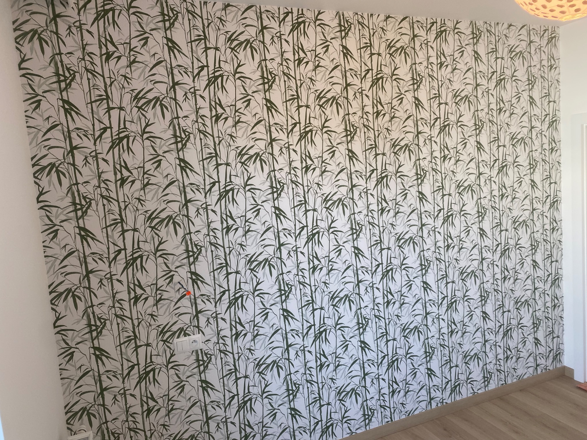 Ściana pokryta tapetą z motywem bambusa, widoczne gniazdko elektryczne i fragment drewnianej podłogi.