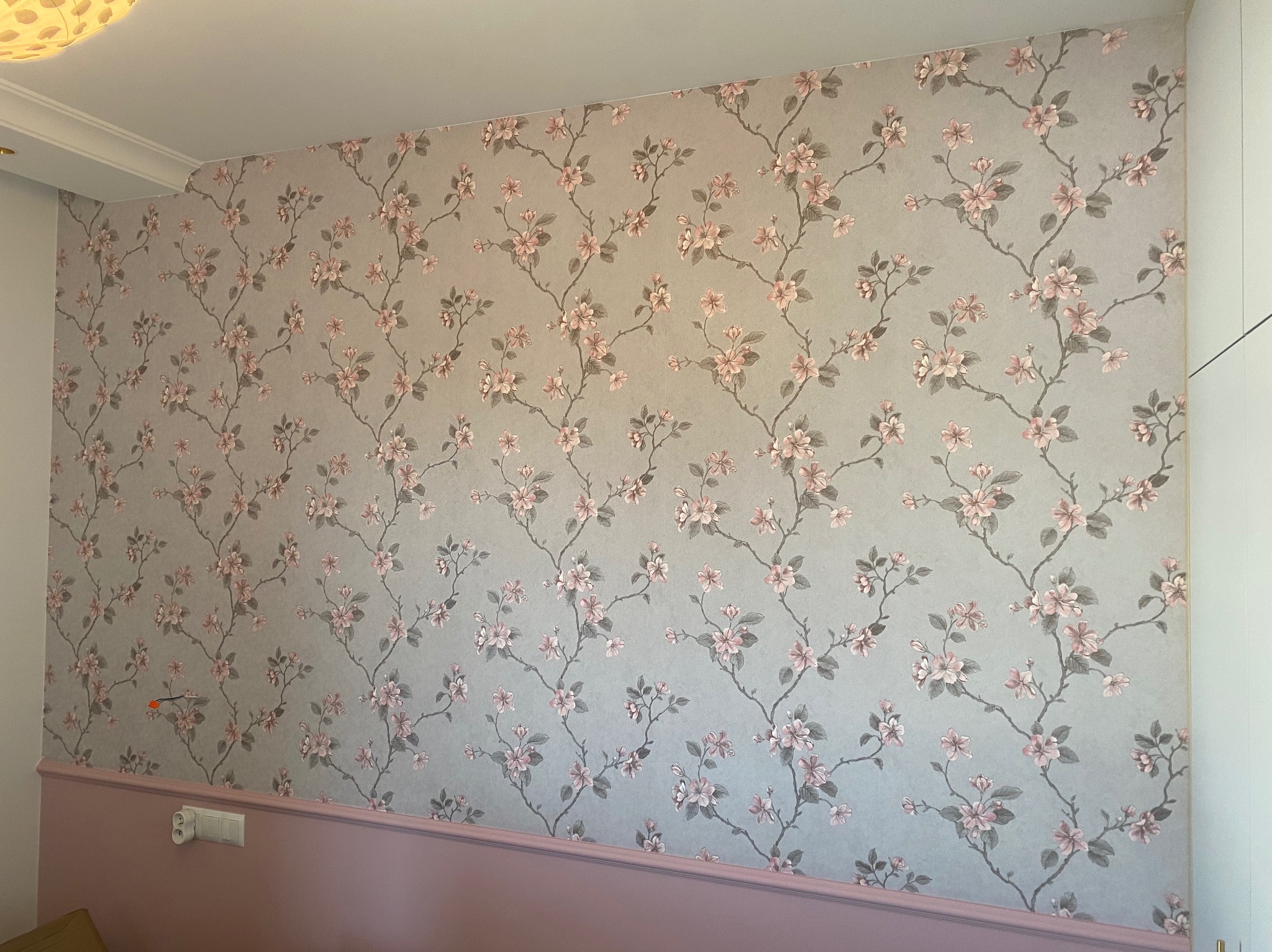 Ściana w Poznaniu wykończona tapetą w kwiaty, z dekoracyjną listwą przypodłogową w odcieniu pudrowego różu i widocznym gniazdkiem elektrycznym.