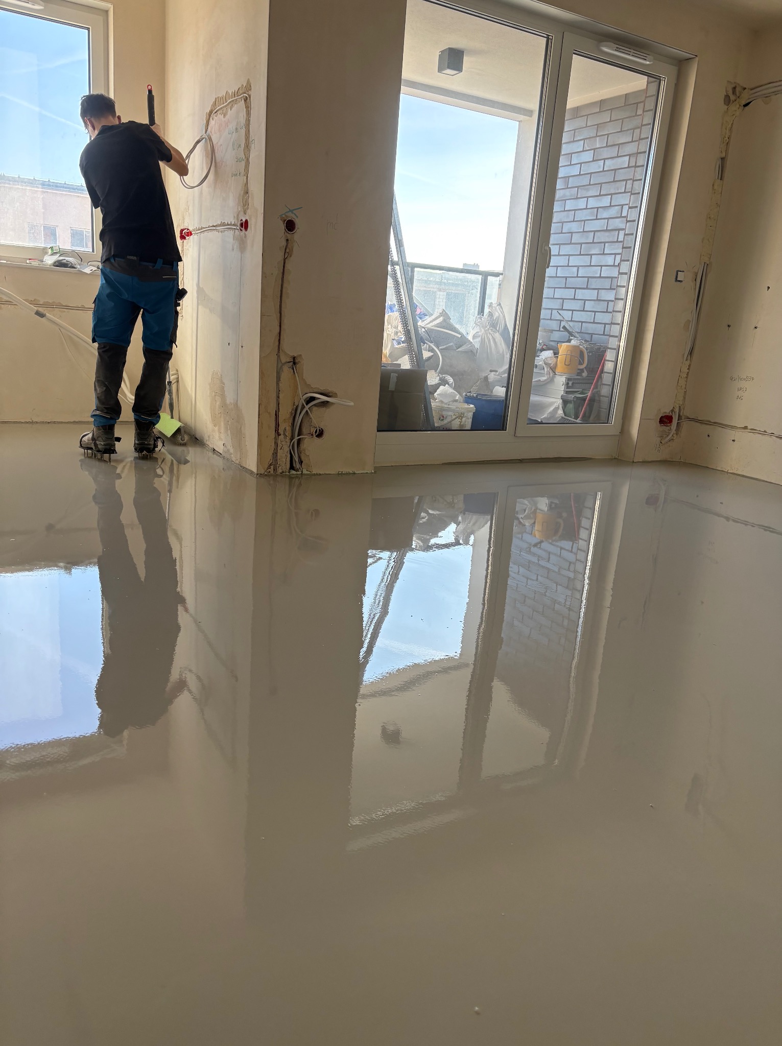 Wylewka samopoziomująca w jasnym pomieszczeniu z dużym oknem balkonowym. Pracownik w butach z kolcami rozprowadza masę. Widoczne instalacje elektryczne w ścianach.