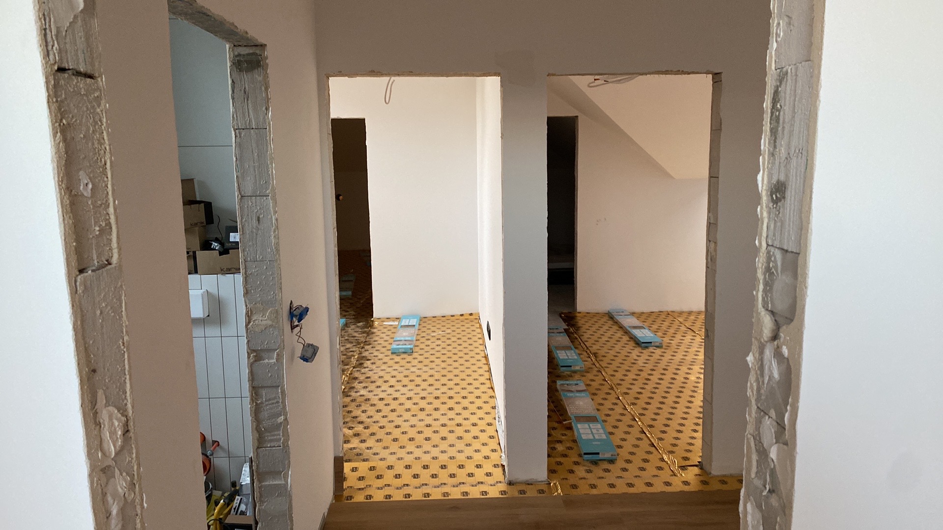 Przygotowanie podłoża pod panele w dwóch pomieszczeniach, widoczne przez surowe futryny drzwiowe, z użyciem podkładu i poziomujących podkładek.