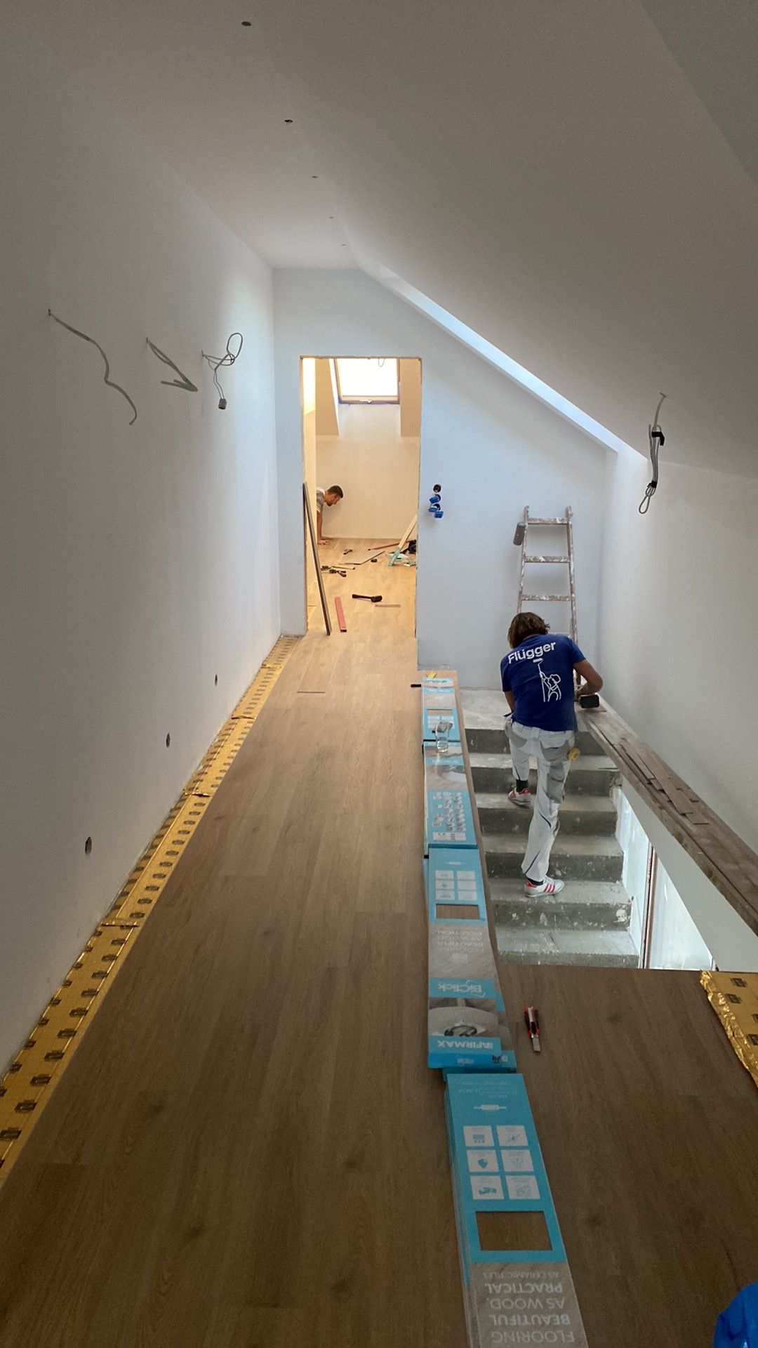 Montaż paneli podłogowych w nowym domu: pracownik w koszulce z logo wchodzi po schodach, widoczne paczki paneli i listwy przypodłogowe wzdłuż ściany.