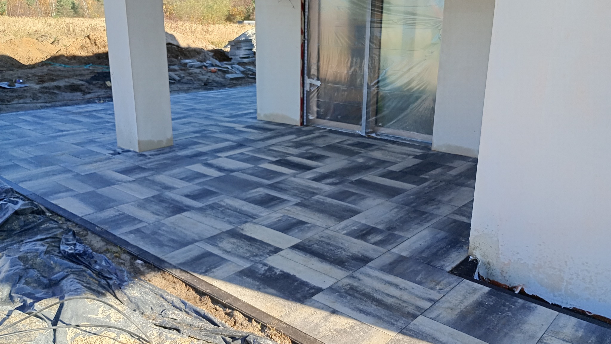 Nowoczesny taras z szarej kostki brukowej ułożonej we wzór. Widoczne kolumny podtrzymujące zadaszenie i przygotowany teren pod dalsze prace.