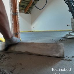 Technobud Henryk Wziątek - Ręczne rozprowadzanie półsuchego jastrychu w pomieszczeniu z widoczną maszyną do zacierania posadzek, listwą dylatacyjną i drewnianą konstrukcją dachu w tle.