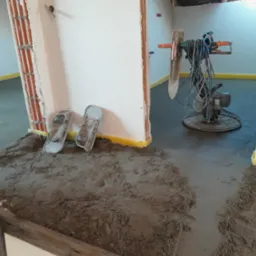 Świeżo wylana, mokra posadzka jastrychowa w trakcie wygładzania maszyną, widoczne robocze klapki i fragment ściany z cegły.