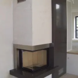 Nowoczesny kominek narożny z wkładem szklanym, obudowany płytami w odcieniach szarości i bieli, z metalowym wykończeniem. Widoczny fragment wentylacji.