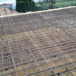 Siatka zbrojeniowa ułożona na deskowaniu przed betonowaniem, widoczne dystanse z tworzywa sztucznego, w tle ziemia i fragment budynku.