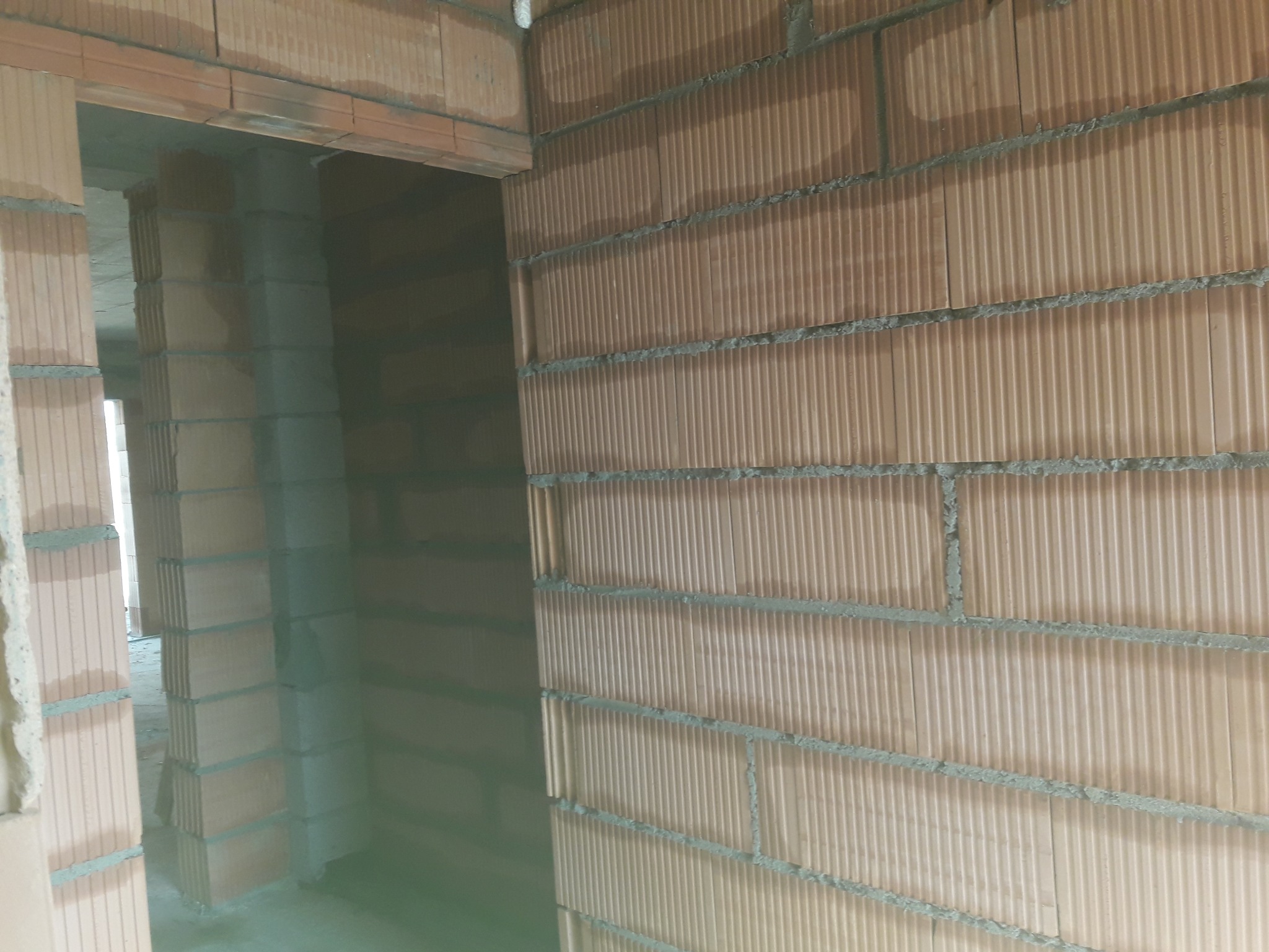 Wnętrze budynku w trakcie budowy, widoczne ściany z pustaków ceramicznych, spoiny z zaprawy, oraz otwór drzwiowy w stanie surowym.