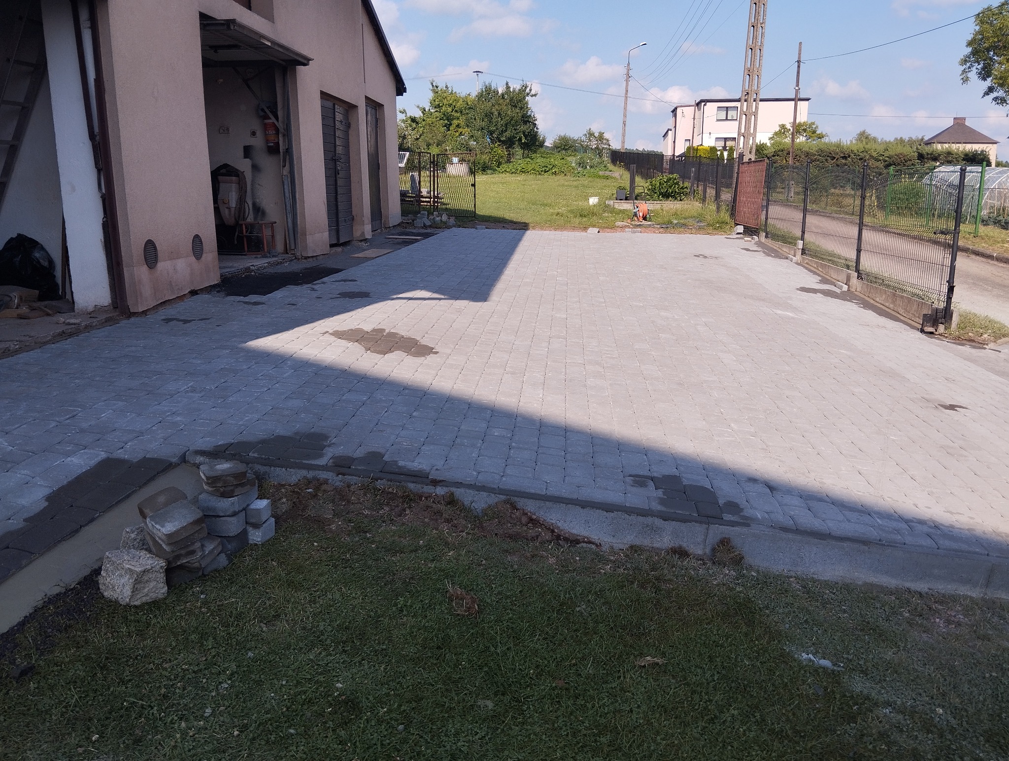 Ułożona szara kostka brukowa na podjeździe z widocznymi mokrymi plamami, obok sterta kostki do wykończenia, w tle budynek gospodarczy i brama wjazdowa.