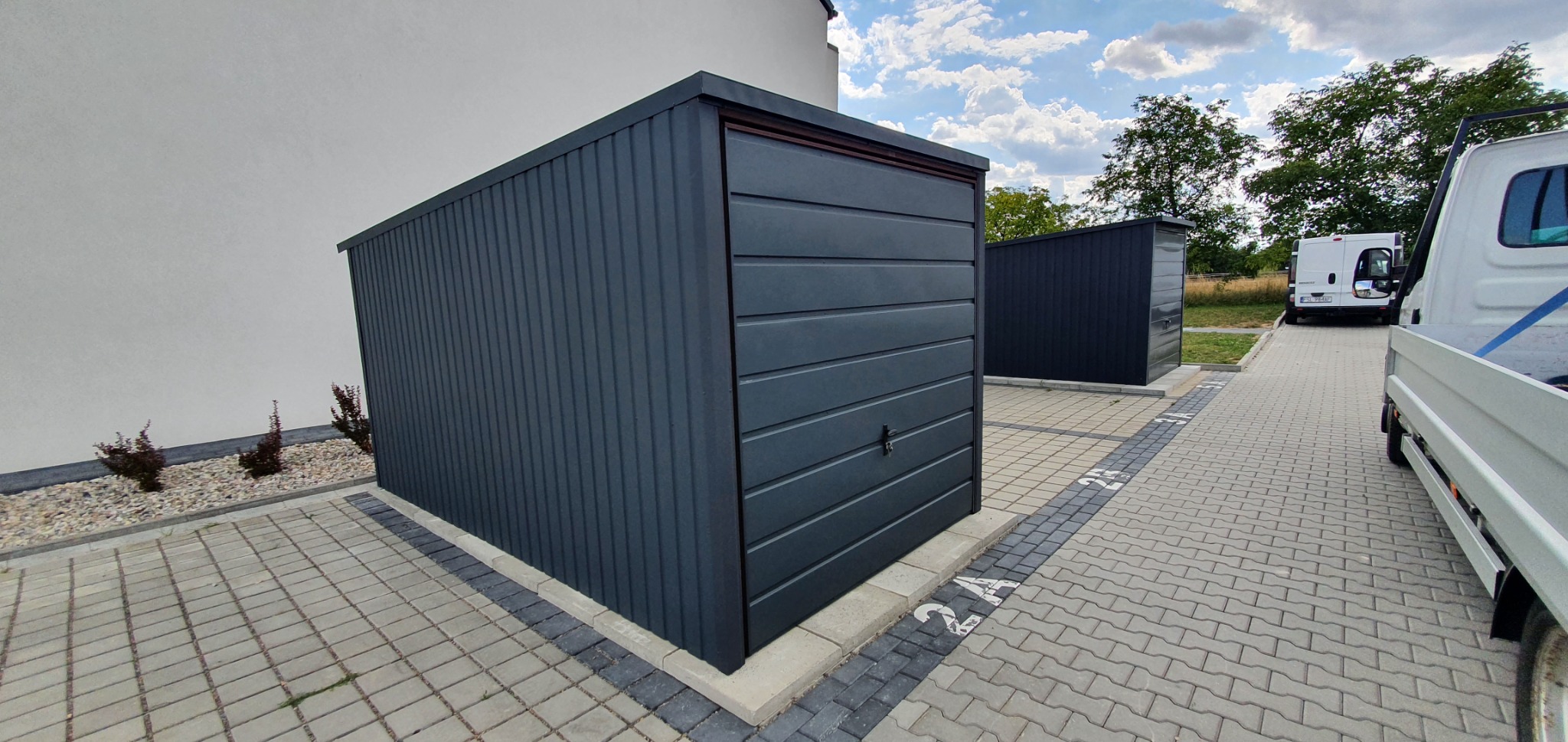 Dwa ciemnoszare garaże blaszane na kostce brukowej, z widocznym fragmentem białej ściany budynku i samochodem dostawczym w tle.